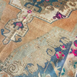 May include: A woven rug featuring a tan background with intricate geometric and floral patterns in shades of dark blue, muted green, and vibrant magenta. The design includes symmetrical motifs and a detailed border with repeating floral elements.