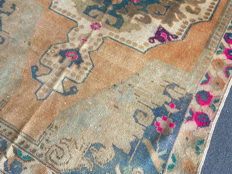 May include: A woven rug featuring a tan background with intricate geometric and floral patterns in shades of dark blue, muted green, and vibrant magenta. The design includes symmetrical motifs and a detailed border with repeating floral elements.