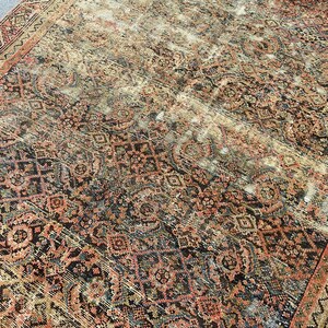 May include: An antique rug with a distressed, faded appearance. The rug features a complex pattern of geometric and floral designs in shades of red, black, and beige. The edges are bordered with a detailed pattern. The rug appears to be worn.