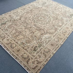 May include: A large, rectangular, beige rug with an intricate floral and geometric pattern. The rug features a central medallion design and a border with repeating motifs in shades of brown and green. The rug's aged appearance suggests it is vintage or antique.