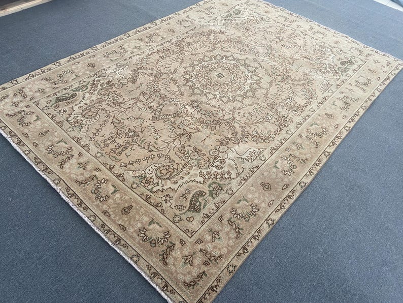 May include: A large, rectangular, beige rug with an intricate floral and geometric pattern. The rug features a central medallion design and a border with repeating motifs in shades of brown and green. The rug's aged appearance suggests it is vintage or antique.