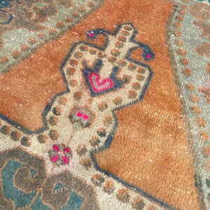 May include: A woven rug featuring a central orange field with a symmetrical geometric medallion in cream, dark blue, and vibrant pink. The design includes intricate borders and repeating patterns in muted teal, brown, and beige tones throughout.