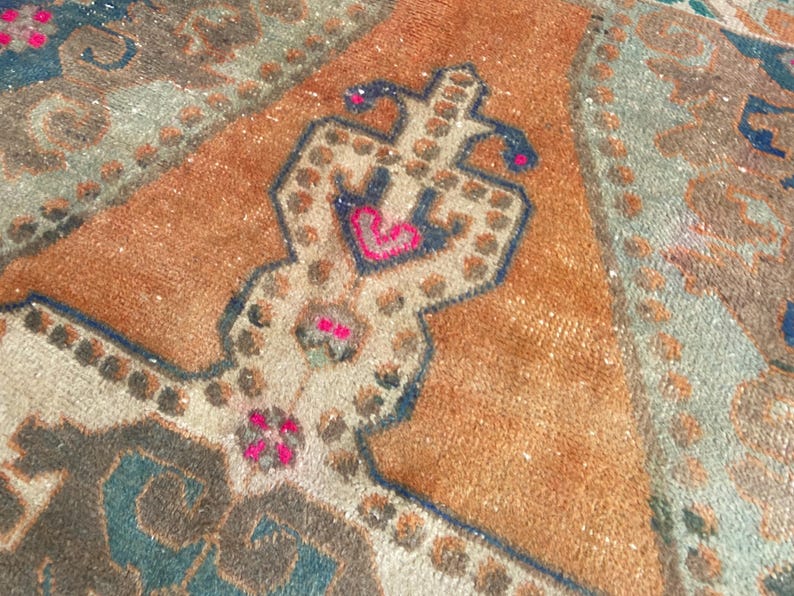 May include: A woven rug featuring a central orange field with a symmetrical geometric medallion in cream, dark blue, and vibrant pink. The design includes intricate borders and repeating patterns in muted teal, brown, and beige tones throughout.