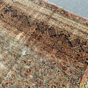May include: An antique Persian rug with a distressed, faded appearance. The rug features a complex, repeating geometric pattern in shades of red, navy, beige, and brown. The rug's edges have a decorative border.