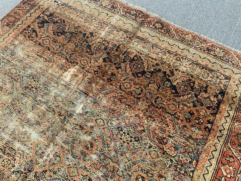 May include: An antique Persian rug with a distressed, faded appearance. The rug features a complex, repeating geometric pattern in shades of red, navy, beige, and brown. The rug's edges have a decorative border.