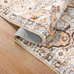 Pode incluir: Tapete enrolado com um padrão floral ornamentado em creme, castanho e cinza. A borda do tapete é visível, revelando uma borda bege e um verso cinza texturizado. O tapete está colocado sobre uma superfície de madeira clara.