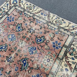 May include: A section of a vintage rug with a faded, distressed appearance. The rug features a salmon-colored central field with floral patterns in blue, teal, and cream. The border is cream with floral and geometric designs.