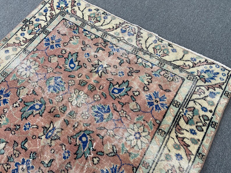 May include: A section of a vintage rug with a faded, distressed appearance. The rug features a salmon-colored central field with floral patterns in blue, teal, and cream. The border is cream with floral and geometric designs.