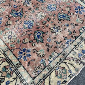 May include: A section of a vintage rug with a faded, distressed appearance. The rug features a central field of muted rose color with floral patterns in blue, teal, and cream. The border is cream with a black outline.