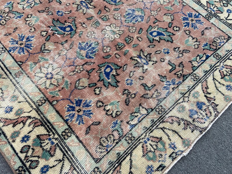 May include: A section of a vintage rug with a faded, distressed appearance. The rug features a central field of muted rose color with floral patterns in blue, teal, and cream. The border is cream with a black outline.