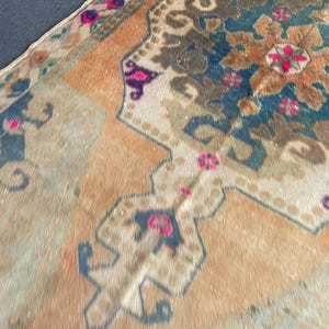 May include: A woven rug featuring a large central medallion in deep blue and brown tones, surrounded by a light orange field. The design includes intricate floral motifs and geometric borders accented with vibrant pink and purple details.