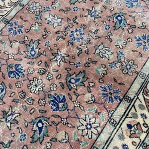 May include: Close-up of a vintage rug with a distressed, faded appearance. The rug features a repeating floral pattern in shades of pink, blue, and cream. The border has a cream background with floral and geometric designs. The rug's texture is visible.