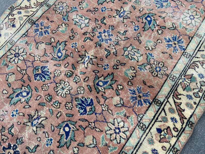 May include: Close-up of a vintage rug with a distressed, faded appearance. The rug features a repeating floral pattern in shades of pink, blue, and cream. The border has a cream background with floral and geometric designs. The rug's texture is visible.