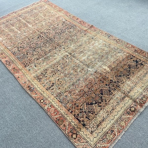 May include: An antique Persian rug with a distressed, vintage appearance. The rug features a central field of muted browns, tans, and blues, with a detailed border in shades of red and beige. The intricate patterns and faded colors give it a classic, aged look.