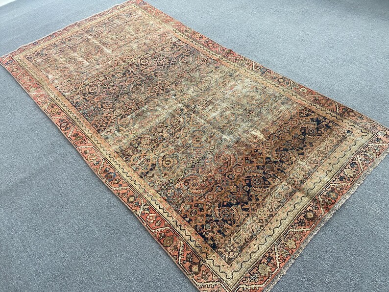 May include: An antique Persian rug with a distressed, vintage appearance. The rug features a central field of muted browns, tans, and blues, with a detailed border in shades of red and beige. The intricate patterns and faded colors give it a classic, aged look.