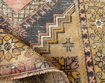 May include: A woven rug featuring a complex geometric pattern with a color palette of muted pink, charcoal gray, mustard yellow, and cream. The design includes zigzag lines, floral motifs, and a fringed edge where the rug is folded over itself.