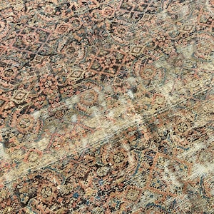 May include: Close-up of a distressed, vintage rug with an intricate, geometric pattern. The rug features a blend of colors, including faded reds, browns, blues, and creams, creating a worn, antique aesthetic. The design includes floral and diamond shapes.