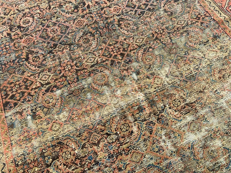 May include: Close-up of a distressed, vintage rug with an intricate, geometric pattern. The rug features a blend of colors, including faded reds, browns, blues, and creams, creating a worn, antique aesthetic. The design includes floral and diamond shapes.