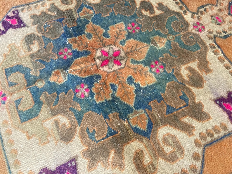 May include: A woven rug featuring a central floral medallion in shades of orange, deep blue, and brown. The design includes intricate geometric patterns and small, vibrant pink flower accents set against a light beige and orange background.