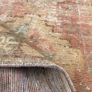 May include: A close-up of a vintage rug with a distressed, faded appearance. The rug features a blend of colors, including peach, beige, and muted blue, creating a warm, inviting aesthetic. The edges are slightly frayed, adding to its antique charm.