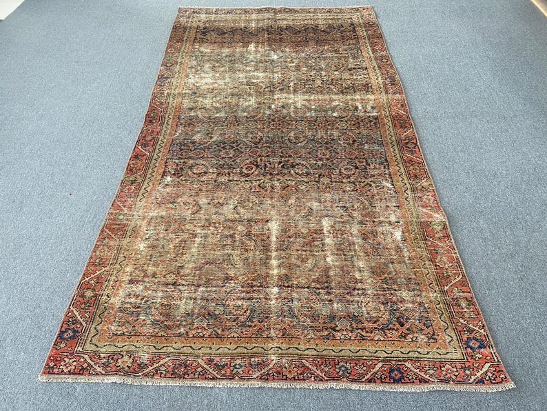 May include: An antique, rectangular rug with a distressed, faded appearance. The rug features a central field of muted colors, including beige, brown, and hints of blue. A detailed border in shades of red and navy frames the rug.