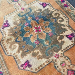 May include: A rectangular woven rug featuring a central medallion in shades of deep blue, brown, and cream. The background is a warm orange hue, accented with intricate floral patterns in vibrant pink, dark purple, and muted green throughout.
