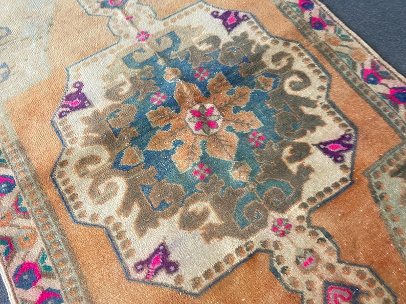May include: A rectangular woven rug featuring a central medallion in shades of deep blue, brown, and cream. The background is a warm orange hue, accented with intricate floral patterns in vibrant pink, dark purple, and muted green throughout.