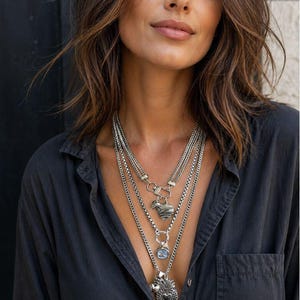 May include: A multi-layered silver-toned chain necklace featuring three distinct pendants: a textured heart, a square-cut clear crystal, and an ornate sacred heart charm. The necklace is worn over a dark charcoal button-down shirt with a chest pocket.