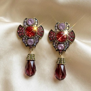May include: A pair of dangle earrings featuring a flower-shaped stud with a central red crystal, surrounded by smaller lavender and red crystals set in antique gold-tone metal. A faceted, teardrop-shaped dark purple crystal hangs from each stud.