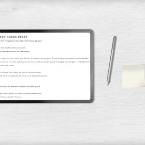 May include: A digital tablet with the text "DEEP FOCUS RESET" and other German text, a silver stylus, and a small yellow sticky note on a light wood-grain surface. The tablet's screen displays text about focus and productivity.