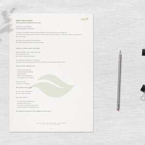 May include: A white sheet of paper with German text, a gray pencil, and two black binder clips on a light wood-grain surface. The paper has the title "DEEP FOCUS RESET" and a leaf-shaped logo. The pencil is sharpened and ready for use.