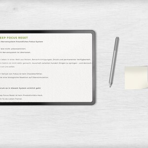 May include: A digital tablet with the text "DEEP FOCUS RESET" and other German text. A silver stylus and a small yellow sticky note are to the right of the tablet. The background is a light wood grain.