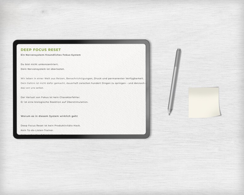 May include: A digital tablet with the text "DEEP FOCUS RESET" and other German text. A silver stylus and a small yellow sticky note are to the right of the tablet. The background is a light wood grain.