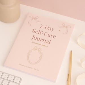 May include: A pink 7-Day Self-Care Journal with gold accents. The journal cover features the text "7-Day Self-Care Journal" and decorative bows. A gold pen rests beside the journal. Designed by Sophie's Edit Co.