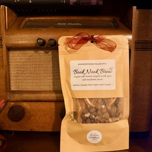May include: A brown paper bag of "Book Nook Brew" stovetop home fragrance, tied with a red ribbon, sits in front of a vintage radio. The bag's label lists ingredients like coffee bean, chamomile, and vanilla.