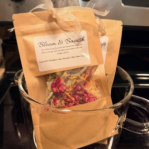 May include: Two small brown paper pouches labeled "Bloom & Banish" filled with dried herbs and flowers, tied with a cream ribbon, inside a clear glass mug. The label lists ingredients: Lemon Peel, Eucalyptus, Sage, Rosemary, Mint, Rose, and Calendula.