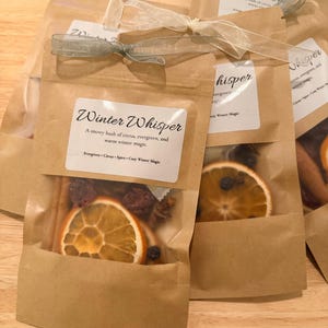 Può includere: Diverse buste di carta marrone con finestre trasparenti, ciascuna contenente fette d'arancia essiccate, bacche e spezie. Le buste sono legate con un nastro. L'etichetta recita "Winter Whisper".