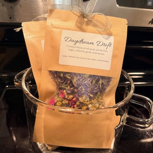 May include: Two small, tan paper pouches filled with dried flowers and herbs, tied with a silver ribbon, sit inside a clear glass mug. The pouches have a label that reads "Daydream Drift" and lists ingredients. The mug is on a dark surface.