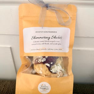 May include: A tan pouch of stovetop home fragrance labeled Shimmering Shores, featuring a clear window revealing dried botanicals. The pouch is tied with a grey ribbon and displays the text The Wishing Wildflower at the bottom center.