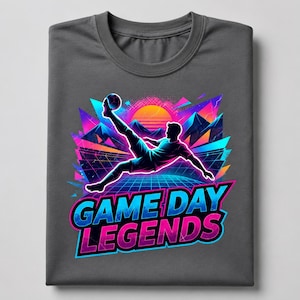 Op de afbeelding: Een antracietgrijs t-shirt met een levendige afbeelding van een voetballer die tegen een bal schopt. Het ontwerp bevat een neonkleurige zonsondergang, bergsilhouetten en een rasterpatroon. De tekst "GAME DAY LEGENDS" is gedrukt in dikke, kleurrijke letters.