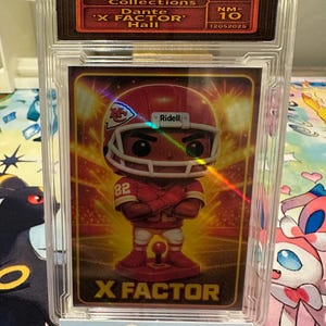 May include: A graded trading card featuring a football player in a red uniform and helmet. The card is encased in a clear plastic holder with the text "2025 TOY POP Collections" and "X FACTOR" visible. The card is graded NM-10.