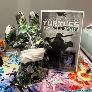 Könnte beinhalten: Eine handförmige Skulptur mit Comic-Design, die eine weiße Maske trägt. Daneben ein Teenage Mutant Ninja Turtles Comic, eine Sammelkarte und eine farbenfrohe Pokémon-Spielmatte.