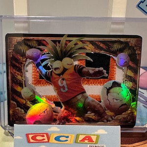 May include: A holographic trading card featuring a cartoon character in an orange jersey with the number 9. The character is throwing several balls. The card is encased in a clear protective sleeve.