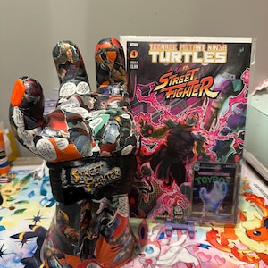 Puede incluir: Una escultura en forma de mano decorada con páginas de cómic, con las palabras "Street Fighter" en la muñeca. Junto a ella hay un cómic titulado "Teenage Mutant Ninja Turtles vs Street Fighter". También se ve una tarjeta coleccionable.