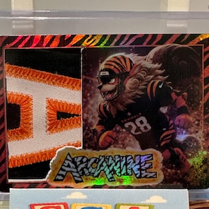 Puede incluir: Tarjeta coleccionable con temática de los Bengals de Cincinnati. Muestra un personaje parecido a un tigre con uniforme de fútbol, con el número 28. La palabra "ARCANE" se muestra en una fuente holográfica colorida. La tarjeta está en un estuche protector.