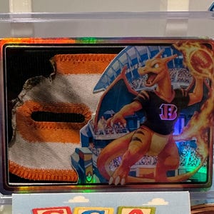 Puede incluir: Una tarjeta coleccionable con una ilustración de Charizard con llamas y un fondo de estadio. La tarjeta está dentro de un soporte protector, con un elemento de tela rasgada en el lado izquierdo. Charizard lleva una camiseta negra con una "B" morada.