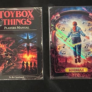 Puede incluir: Dos cartas coleccionables. La carta de la izquierda dice "TOYBOX THINGS PLAYERS MANUAL" con una ilustración de fantasía. La carta de la derecha presenta a una persona rodeada de notas musicales, con la palabra "MADMAX" en la parte inferior. Ambas cartas están en fundas protectoras.