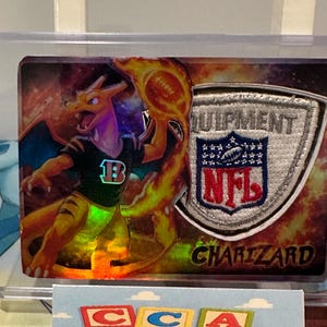 May include: A Charizard trading card with a football theme. The card features a Charizard character in a football uniform, holding a flaming football. The NFL Equipment logo is also visible. The card is encased in a protective sleeve.