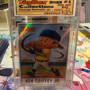 May include: A baseball card encased in a clear protective holder. The card features an illustration of a cartoon baseball player in a yellow jersey with the name "MOELLER" and the text "KEN GRIFFEY JR." The card is labeled "ToyBox 2025 #1 Collections".