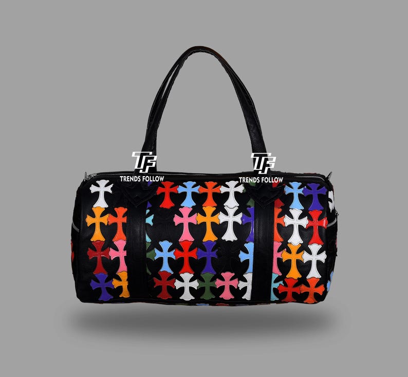 May include: Black duffel bag with a colorful cross pattern. The bag has two black handles and a zipper closure. The crosses are in various colors, including red, blue, orange, and white. The bag has the text "TRENDS FOLLOW" on it.